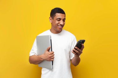 Beyaz tişörtlü genç Afro-Amerikan adam dizüstü bilgisayar tutuyor ve sarı izole edilmiş arka planda akıllı telefon kullanıyor, modern aletleri olan adam telefonda mesaj yazıyor.