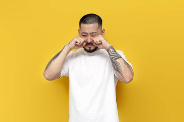 Dövmeli, beyaz tişörtlü, üzgün Asyalı adam sarı izole arka planda ağlıyor, mutsuz, stresli ve depresyonlu Koreli adam.