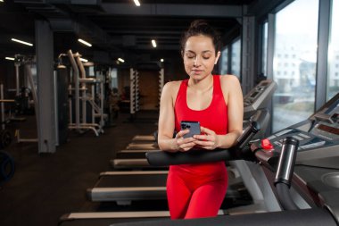 young sporty girl in red sportswear is using smartphone in the gym and typing a message, attractive slim woman is holding the phone in training
