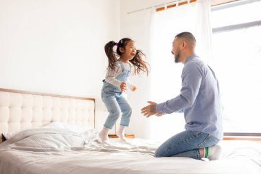 Küçük aktif Asyalı kız babasıyla evde oynuyor, enerjik bir çocuk ailesiyle yatakta zıplıyor ve dans ediyor, Koreli adam kızını kusuyor ve seviniyor.