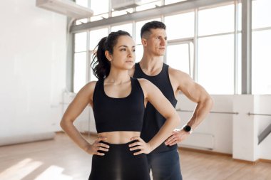 beautiful athletic couple in sportswear stands in the gym in the morning, fitness girl and coach man look to the side at the copy space at workout in bright room