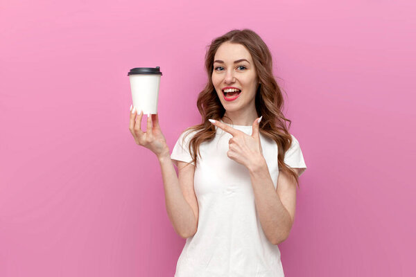 young cute woman with paper cup of coffee on pink isolated background, girl in white t-shirt shows and advertises drink