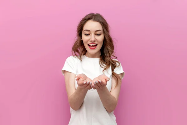 young cute woman in white t-shirt holds empty hands in front of her on pink isolated background, the girl holds nothing in her palms