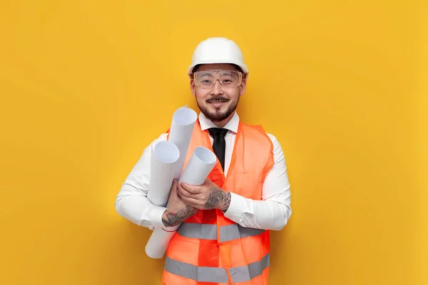 asian builder engineer in uniform holding papers with blueprints on yellow isolated background, male korean in hardhat and work vest, worker architect