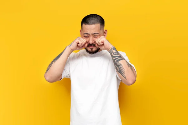 stock image tattooed sad asian man in white t-shirt crying over yellow isolated background, unhappy korean guy in stress and depression