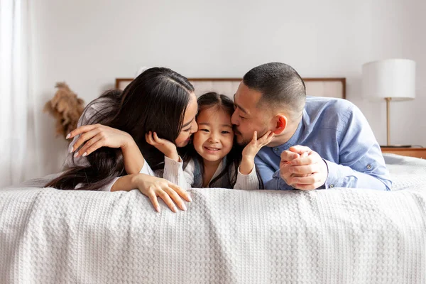 happy young asian family with little daughter lie on bed at home and smile, korean girl together with parents rest at home on bed, mom and dad kiss and love daughter