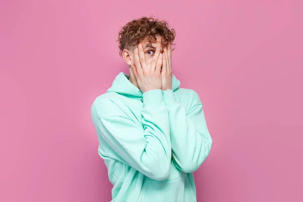 young curly guy covers his face with his hands and peeps through his fingers, man in mint hoodie is afraid and peeps with one eye on pink background
