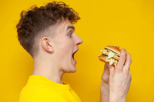 Young Curly Guy Holding Burger His Mouth Open Yellow Background — Stok fotoğraf