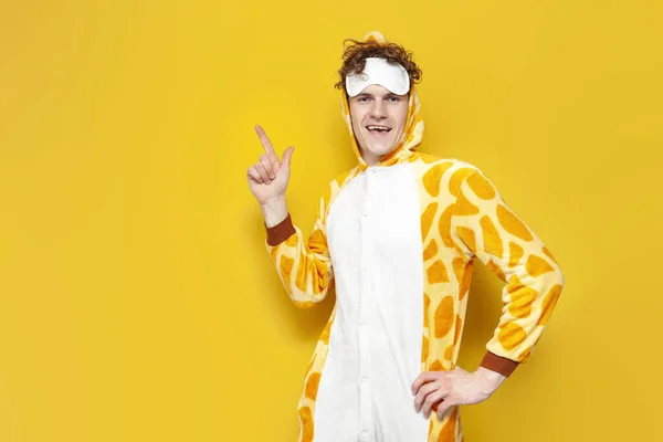 stock image young joyful guy in funny baby giraffe pajamas and sleep mask shows his hand to the side on yellow background, man in animal cosplay clothes advertises copy space