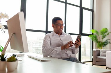 Beyaz gömlekli ve gözlüklü Afro-Amerikan bir adam ofiste bilgisayarın başına oturur ve akıllı telefon kullanır. Ofis çalışanı masaya oturur ve telefona bakar.