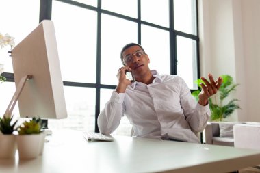 İş kıyafetleri ve gözlüklü Afro-Amerikan bir adam ofiste bilgisayar başında oturmuş telefonla konuşuyor, ofis çalışanı masada oturuyor ve cep telefonu görüşmesi yapıyor.