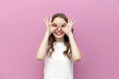 Genç ve neşeli kadın elleriyle maske yapıyor ve pembe izole arkaplandan ona bakıyor. Kız elleriyle gözlük taklit ediyor ve parmaklarından bakıyor.