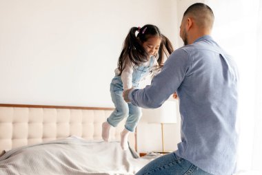 Küçük aktif Asyalı kız babasıyla evde oynuyor, enerjik bir çocuk ailesiyle yatakta zıplıyor ve dans ediyor, Koreli adam kızını kusuyor ve seviniyor.