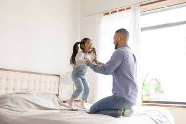 Küçük aktif Asyalı kız babasıyla evde oynuyor, enerjik bir çocuk ailesiyle yatakta zıplıyor ve dans ediyor, Koreli adam kızını kusuyor ve seviniyor.