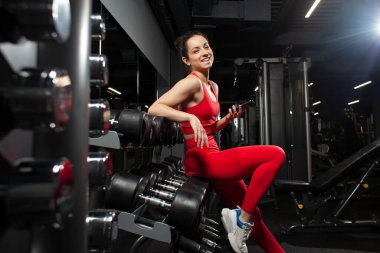 Kırmızı spor kıyafetli atletik bir kadın siyah spor salonunda akıllı telefon kullanıyor. Spor salonunda telefonda mesaj yazan zayıf bir kız. İnternette sohbet eden çekici bir kadın.