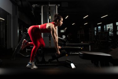 Kırmızı spor kıyafetli atletik bir kadın siyah spor salonunda antrenman yapıyor. İnce bir kız spor salonunda halteri kaldırıyor. Çekici bir kadın koyu renk spor kulübünde vücut geliştirme yapıyor.
