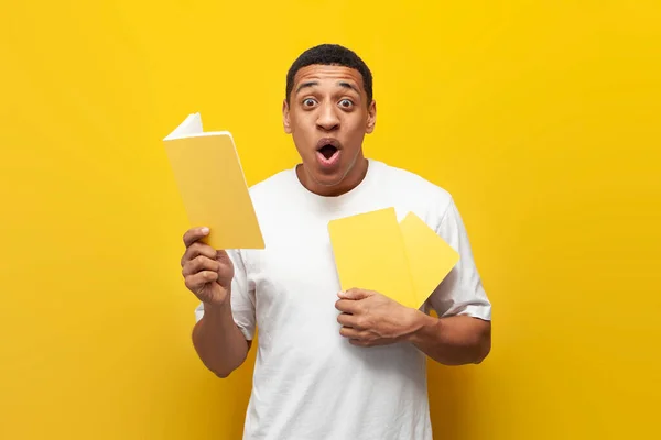 shocked guy african american student in white t-shirt holding yellow book over isolated background and wondering, amazed man reading book