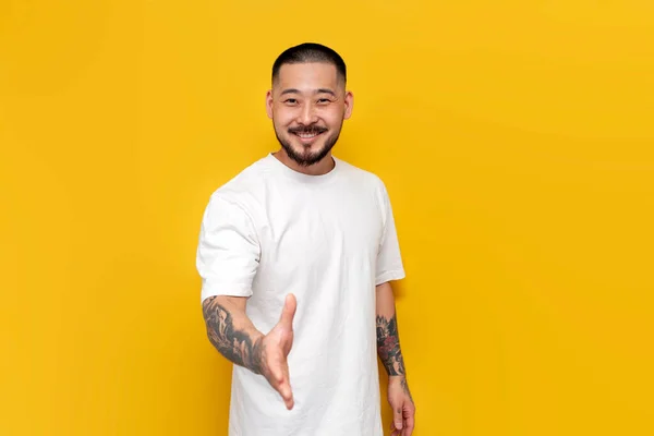 unshaven asian man in white t-shirt says hello and makes handshake gesture on yellow isolated background, adult guy korean greets and extends his hand
