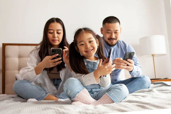 stock image little asian girl with mom and dad use smart phone, korean happy young family with mobile gadgets sit on bed at home and chat online