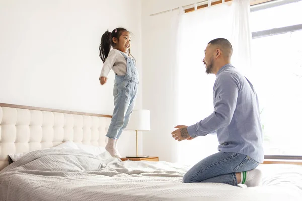 小さなアクティブなアジアの女の子は彼女のお父さんと一緒に家で遊びます 精力的な子供がジャンプし 親とベッドの上で踊ります 韓国の男は彼の娘をスローアップし — ストック写真