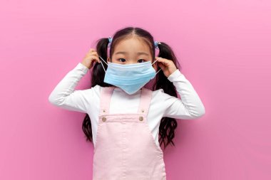 Küçük Asyalı kız pembe soyutlanmış arka plana koruyucu bir maske takıyor, Koreli antiviral maskeli anaokulu çocuğu