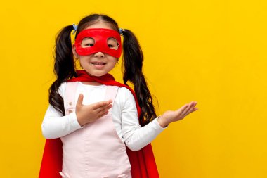 Süpermen kostümü ve maskeli küçük Asyalı kız ellerini kenarda gezdiriyor ve fotokopi alanının reklamını yapıyor. Koreli çocuk sarı izole arka planda gülümsüyor.