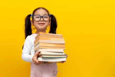 Gözlüklü küçük Asyalı kız kitap tutuyor ve sarı izole edilmiş arka planda gülümsüyor, Koreli akıllı anaokulu öğrenme dersleri alıyor, okula geri dönüyor.