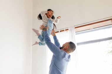 Küçük aktif Asyalı kız babasıyla evde oynuyor, enerjik bir çocuk ailesiyle yatakta zıplıyor ve dans ediyor, Koreli adam kızını kusuyor ve seviniyor.