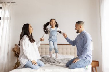 Mutlu Asyalı bir aile kızı ile oynuyor ve evde yatakta eğleniyor. Küçük Koreli kız yatağa atlıyor ve anne ve babayla el ele tutuşuyor.