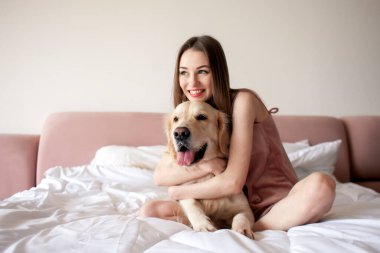 Pijamalı genç tatlı kız yatakta oturur ve Golden Retriever cinsi köpeğe sarılır. Sabahları evcil hayvanıyla evde bir kadın.