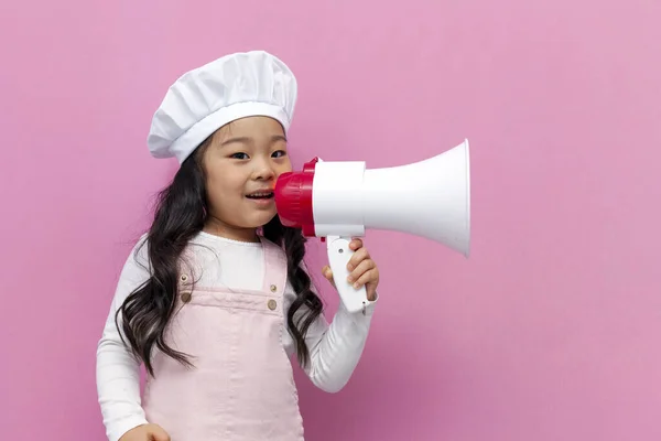 身穿厨师制服的亚洲小女孩在粉色孤立的背景上用扩音器宣布新闻和信息 韩国小孩做饭时尖叫着 对着扩音器说话 — 图库照片