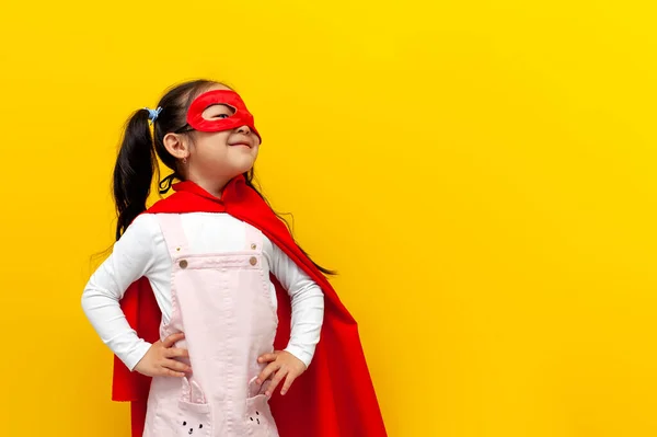 little asian girl in superman costume and mask looks away, korean kid in superhero cape looks at copy space on yellow isolated background