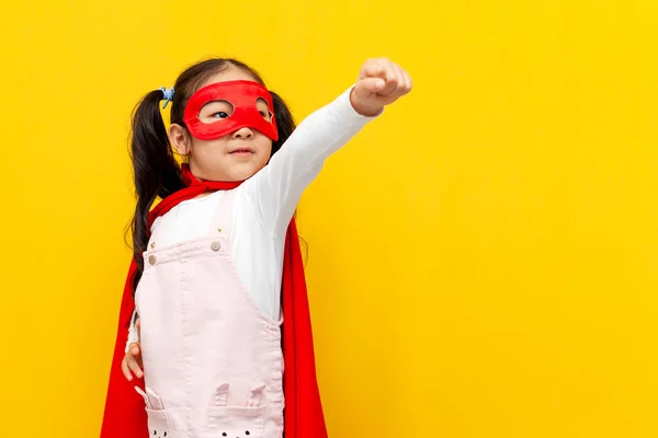 little asian girl in superman costume and mask shows with her hand forward and flies, korean child in superhero cape smiles on yellow isolated background