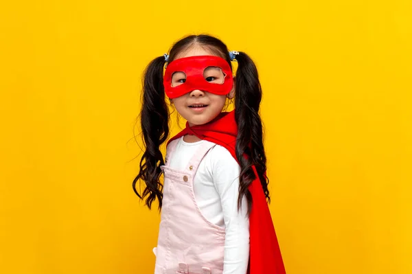 little asian girl in superman costume and mask smiles, korean child in superhero cape on yellow isolated background
