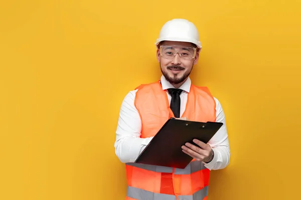Asian civil engineer in uniform writes plan on paper and smiles on yellow isolated background, Korean man in hard hat and work vest works with papers, architect worker