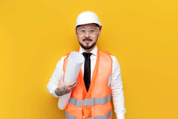 stock image asian foreman in uniform with papers and projects on yellow isolated background, korean civil engineer in hardhat and goggles holding blueprints