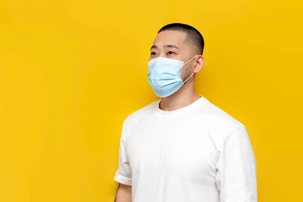 Asian man in medical mask and white football stands on yellow isolated background, Korean guy in protective mask against viruses and flu