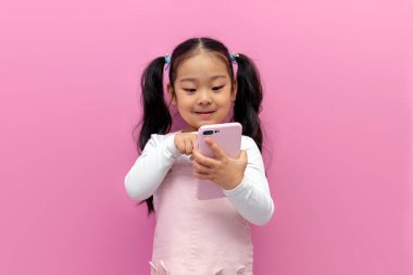 Pembe elbiseli küçük Asyalı kız pembe izole edilmiş arka planda akıllı telefon kullanıyor, Koreli anaokulu çocuğu telefonda yazı yazıyor.
