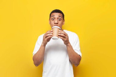 Beyaz tişörtlü genç, şok olmuş Afrikalı Amerikalı adam sarı arka planda kağıt bardakta kahve içiyor ve sürpriz gösteriyor.