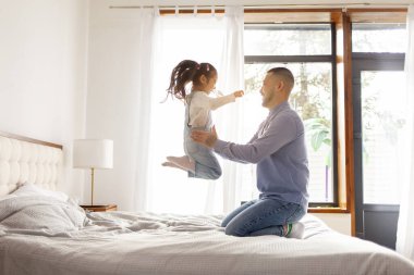 Küçük aktif Asyalı kız babasıyla evde oynuyor, enerjik bir çocuk ailesiyle yatakta zıplıyor ve dans ediyor, Koreli adam kızını kusuyor ve seviniyor.