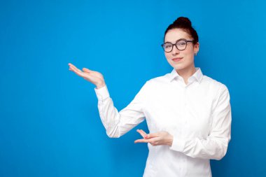 Beyaz tişörtlü ve gözlüklü genç bayan ofis çalışanı ellerini mavi arka planda, kadın sekreter ve muhasebeci renkli izole arka planda fotokopi alanı reklamlarında gösteriyor.