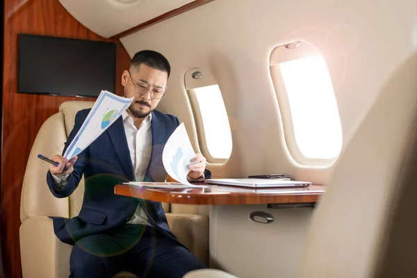 stock image successful asian businessman in suit and glasses sits in private jet and works with documents, korean businessman flies in business class and checks charts and papers