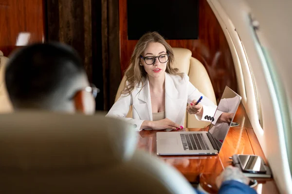 Stock image business meeting on airplane, woman in business clothes sits in private jet and talks with business partner pointing to laptop, business colleagues discuss and plan deal and fly in first class