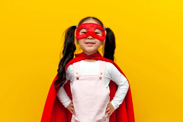 little asian girl in red superman costume and mask, korean child in superhero cape smiling on yellow isolated background
