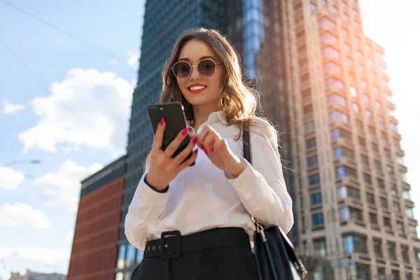 stock image young attractive girl in sunglasses and white shirt is using smartphone on the street in the city and smiling, woman is typing online on the phone