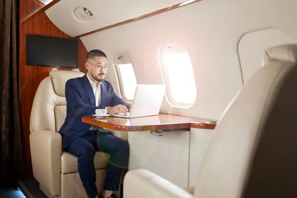 successful asian businessman in suit and glasses sits in private jet and types on laptop, korean entrepreneur in business clothes flies in airplane, luxury lifestyle