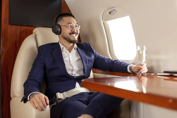 asian businessman in suit sits in private jet drinks champagne and listens to music on headphones, korean entrepreneur flies in airplane and relaxes, luxury lifestyle