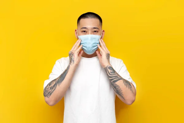 Asian man in medical mask and white football stands on yellow isolated background, Korean guy in protective mask against viruses and flu