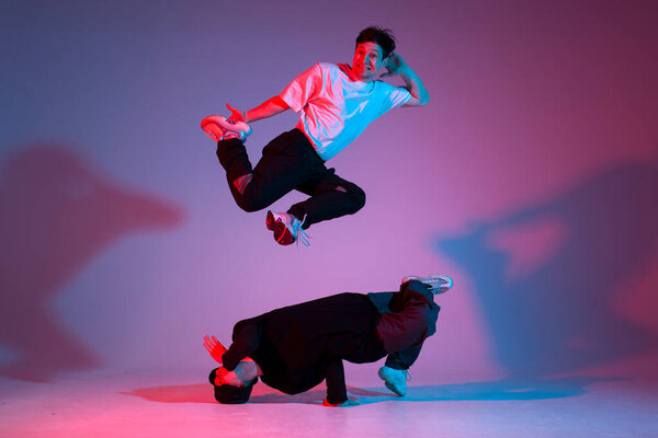 two dancer guys break dance on red blue background and do trick together, male acrobats perform pair dance in neon lighting, hiphop performers