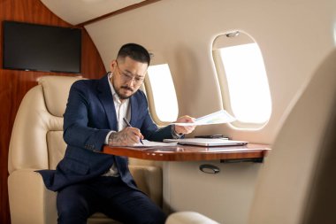 Takım elbiseli ve gözlüklü başarılı Asyalı işadamı özel jette oturuyor ve belgelerle çalışıyor. Koreli finansör, uçakta evrakları ve çizelgeleri kontrol ediyor.